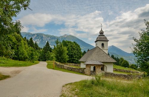 Free Quaint church nestled in lush greenery with mountain backdrop under a bright sky. Stock Photo