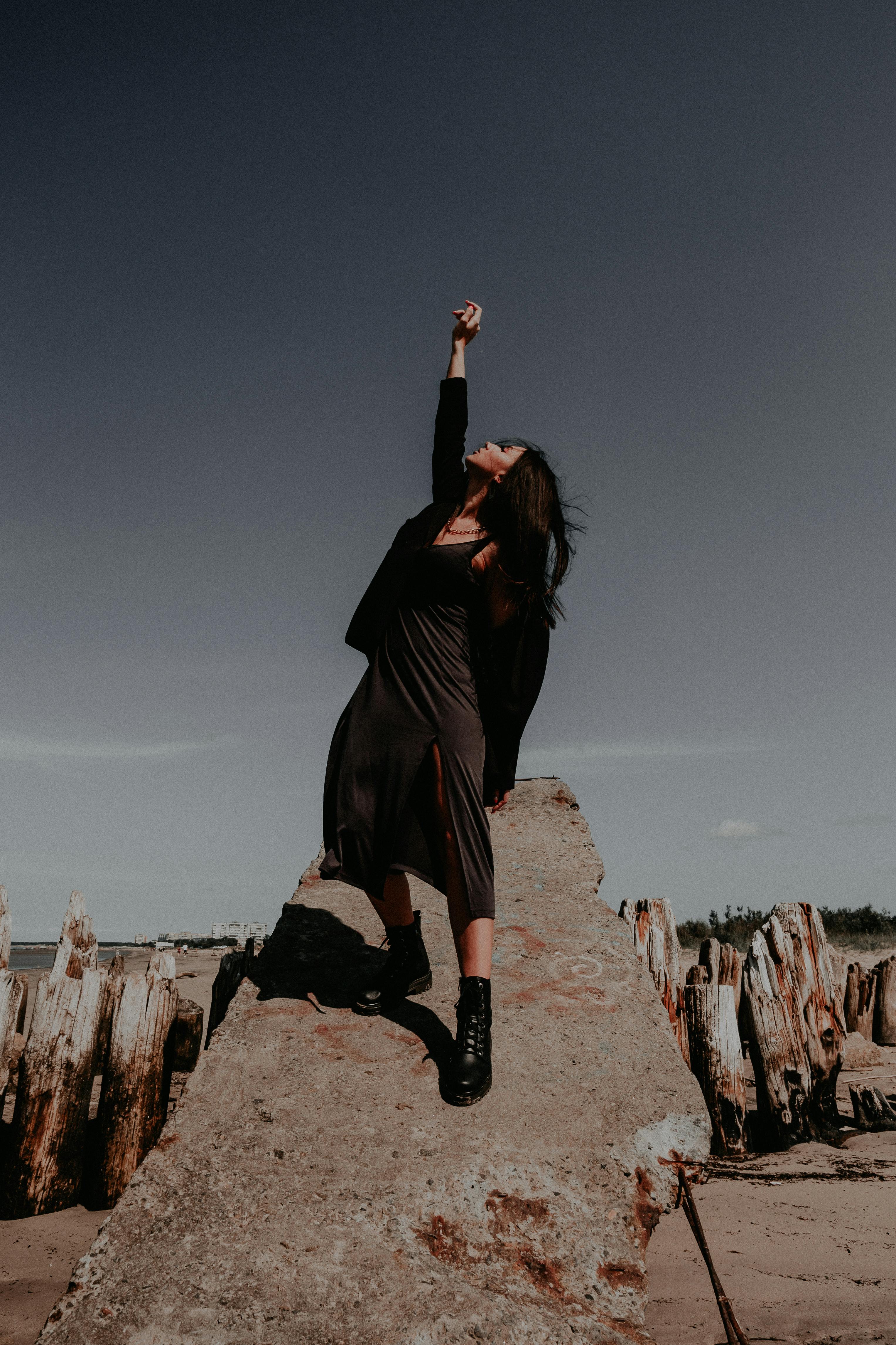 Confident woman in black dress posing with raised hand on a rock outdoors.