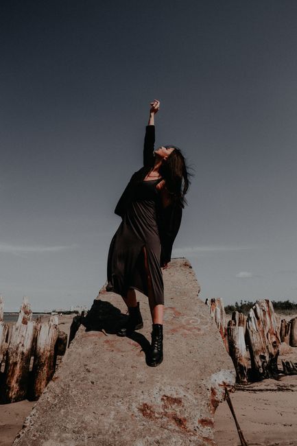 Confident woman in black dress posing with raised hand on a rock outdoors.