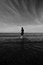 Grayscale Photo of Man in Black Jacket and Pants Walking on Beach