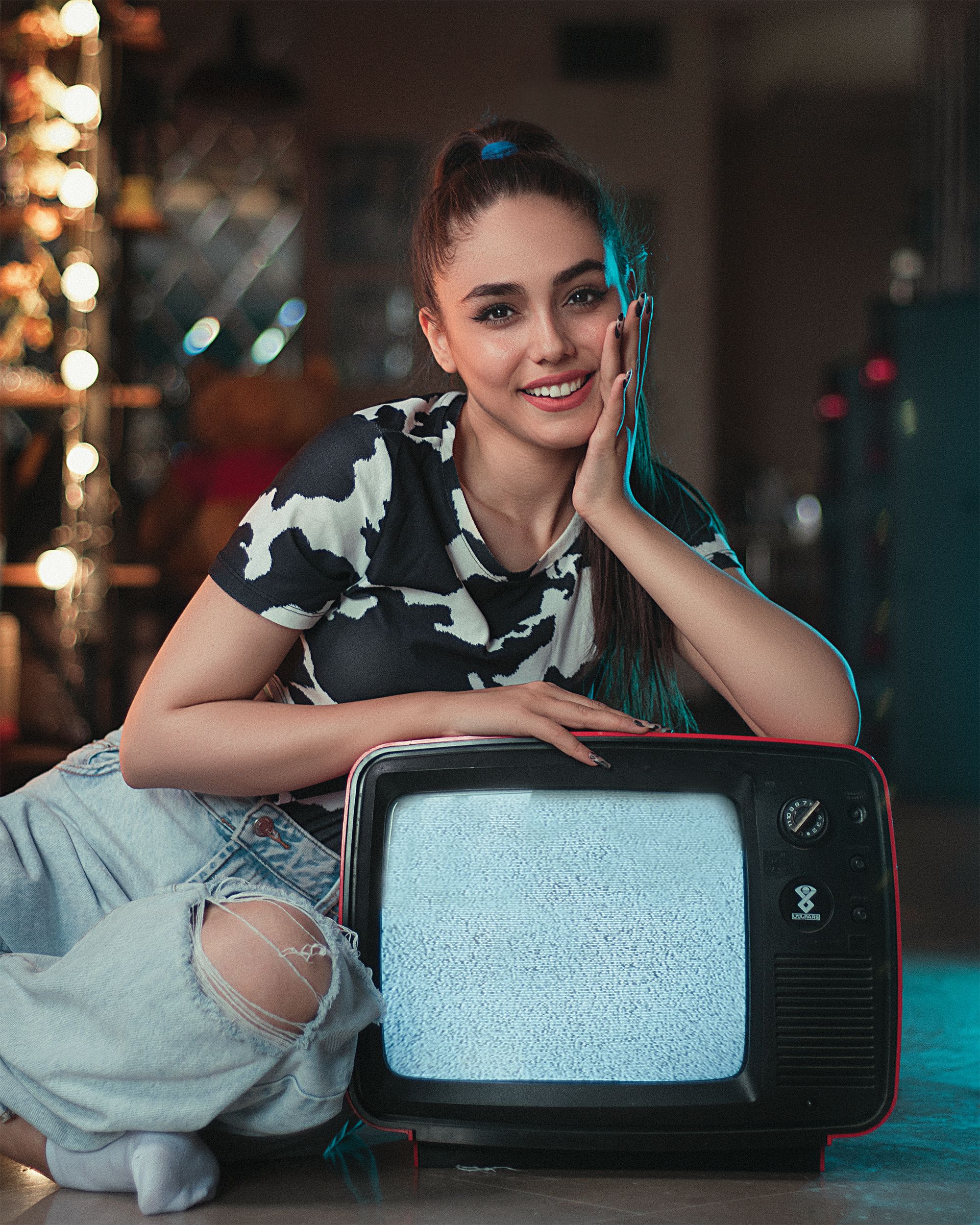 A cheerful woman in ripped jeans posing with a retro TV indoors.