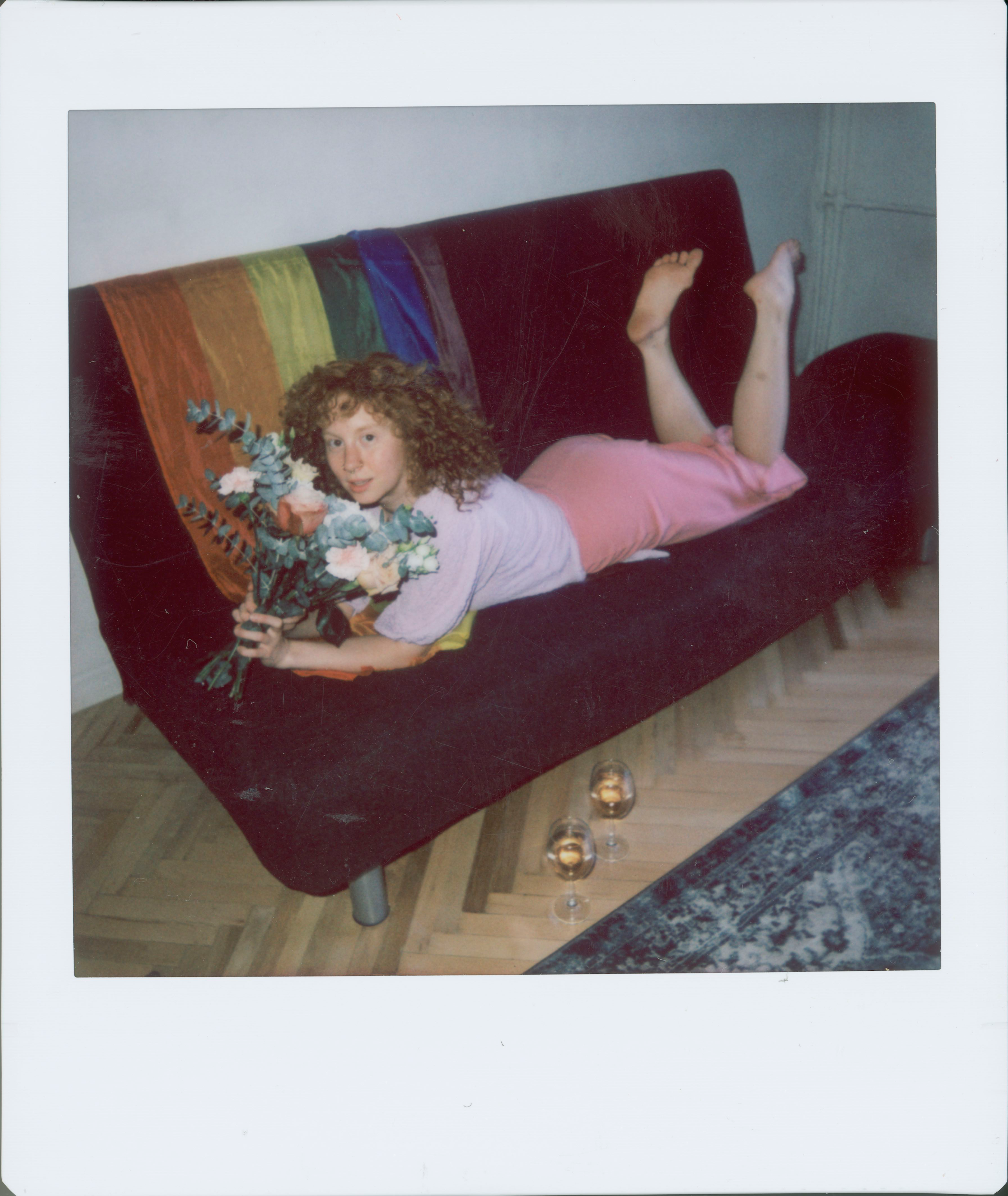 A woman holds a bouquet while lying on a colorful couch indoors, surrounded by candles.