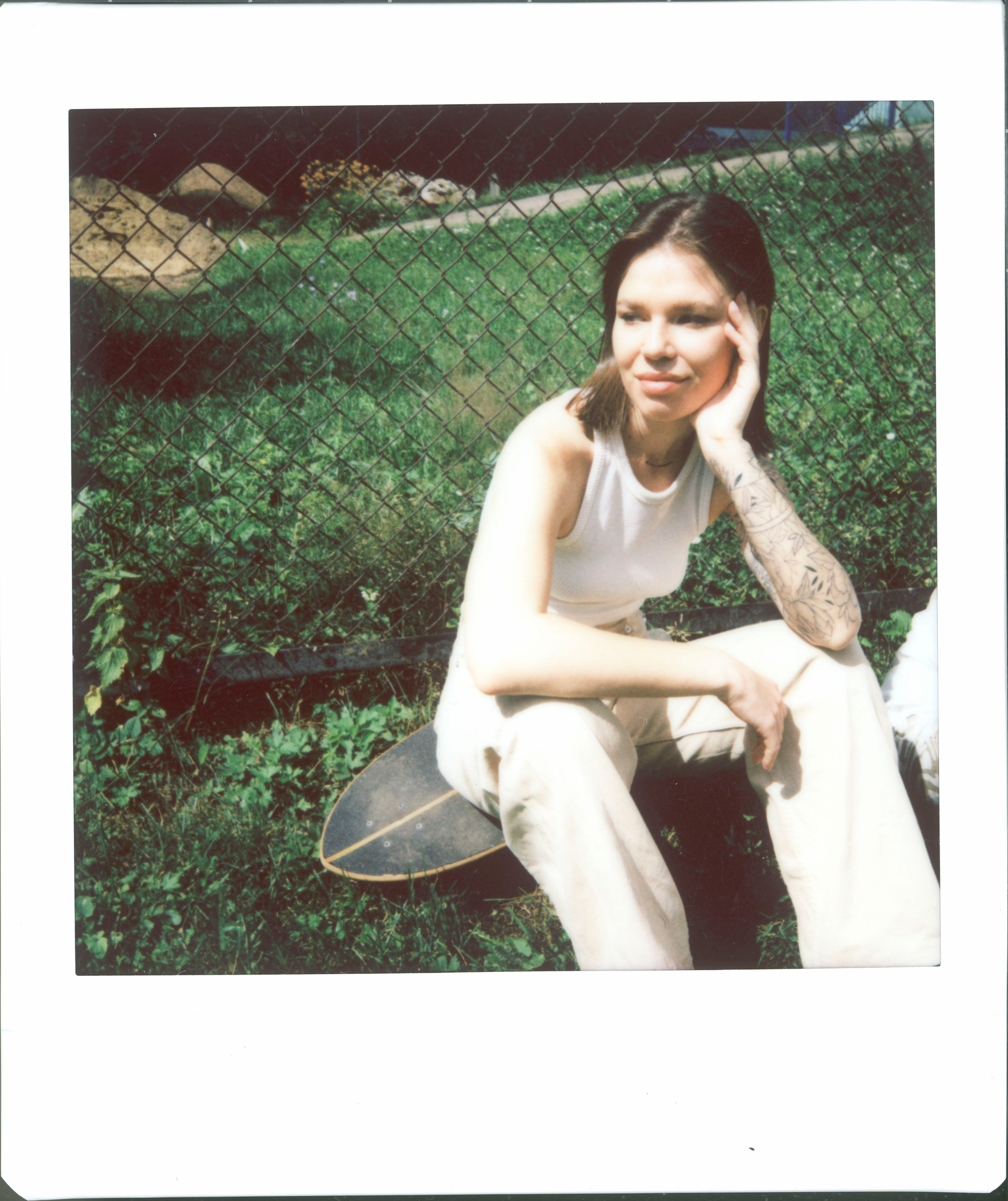 Casual portrait of a young woman sitting on a skateboard in a grassy outdoor setting.