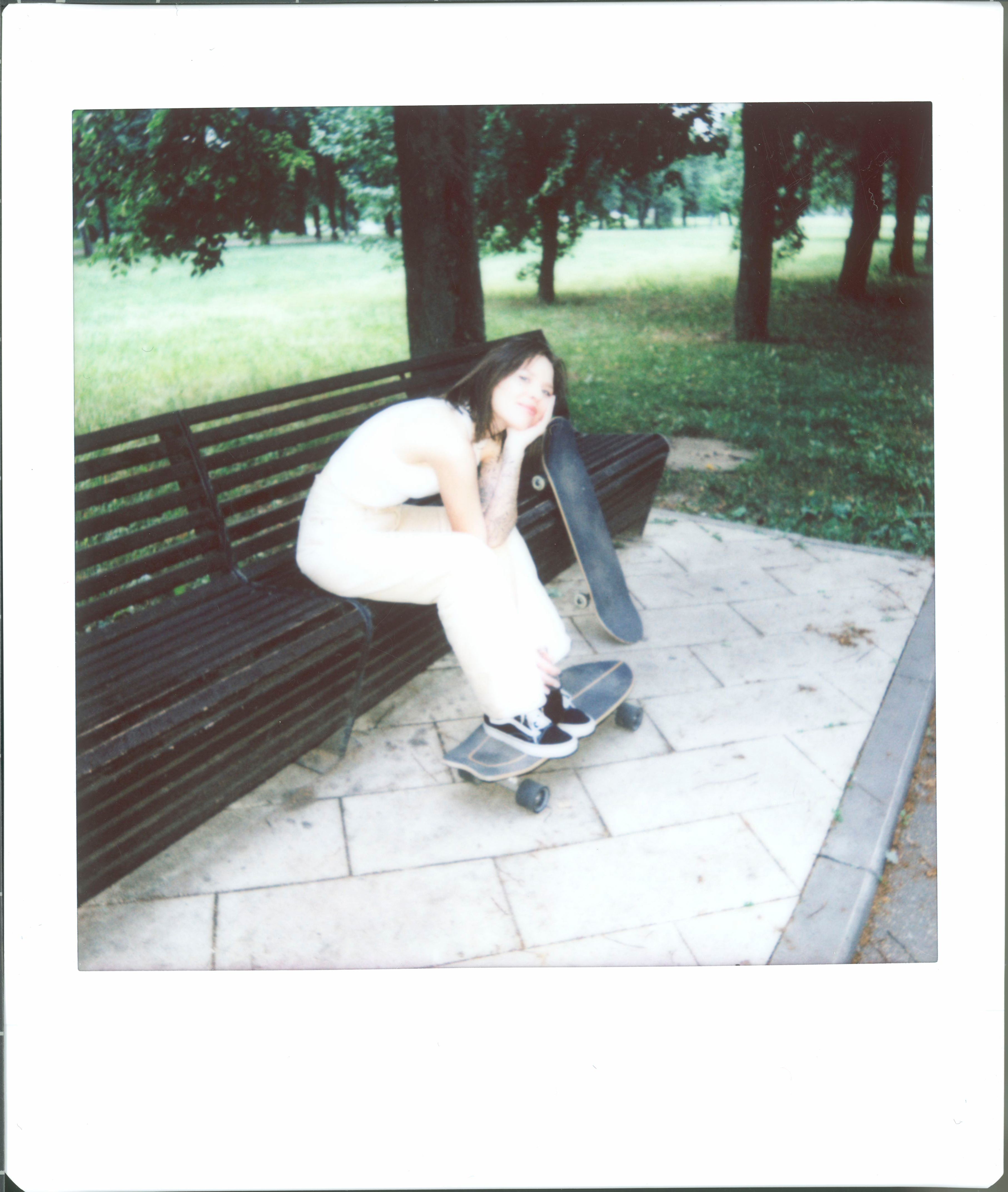 A stylish woman rests on a park bench with a skateboard during a sunny day.