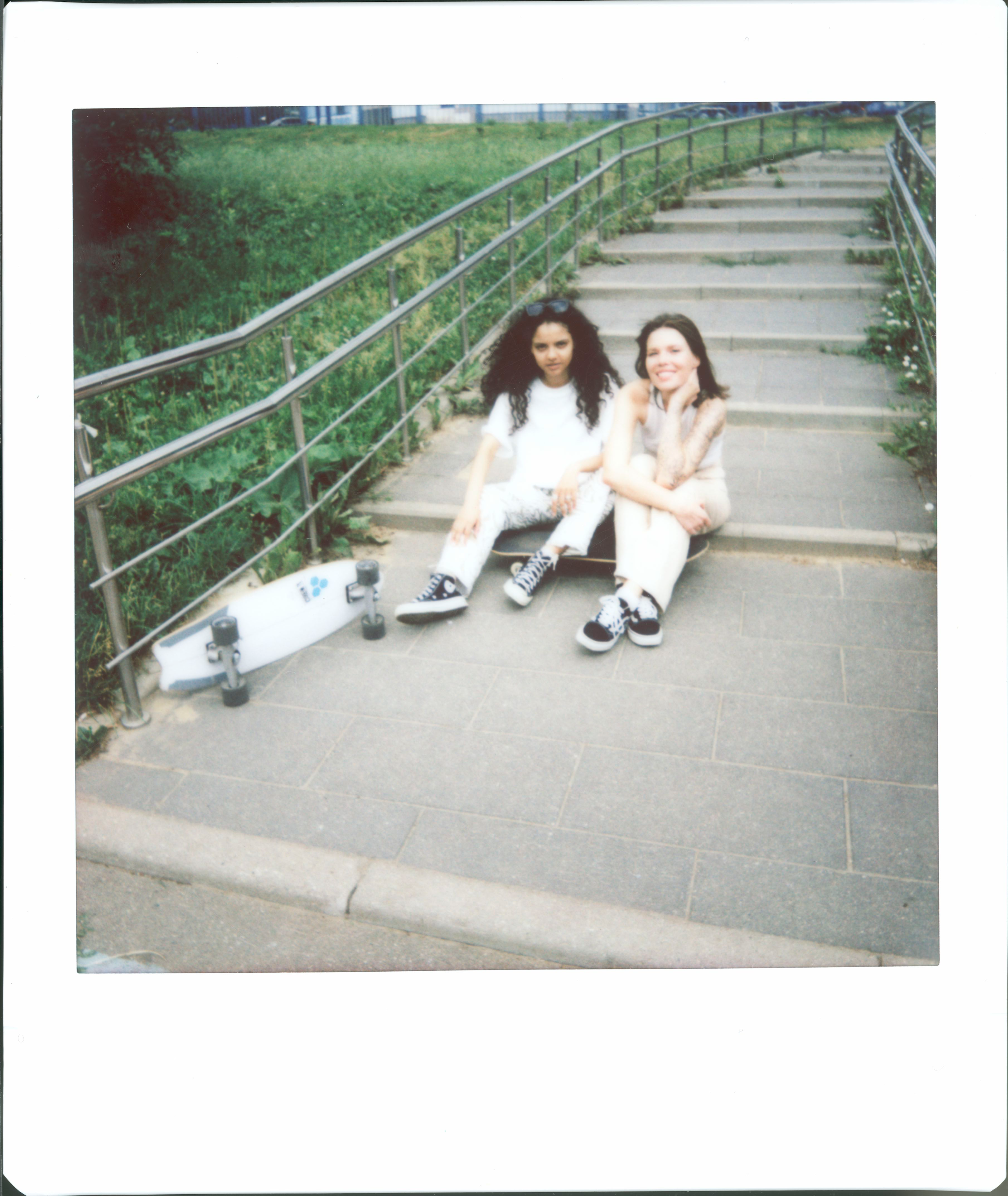 Two skater girls sit on outdoor stairs with skateboards, enjoying a summer day.