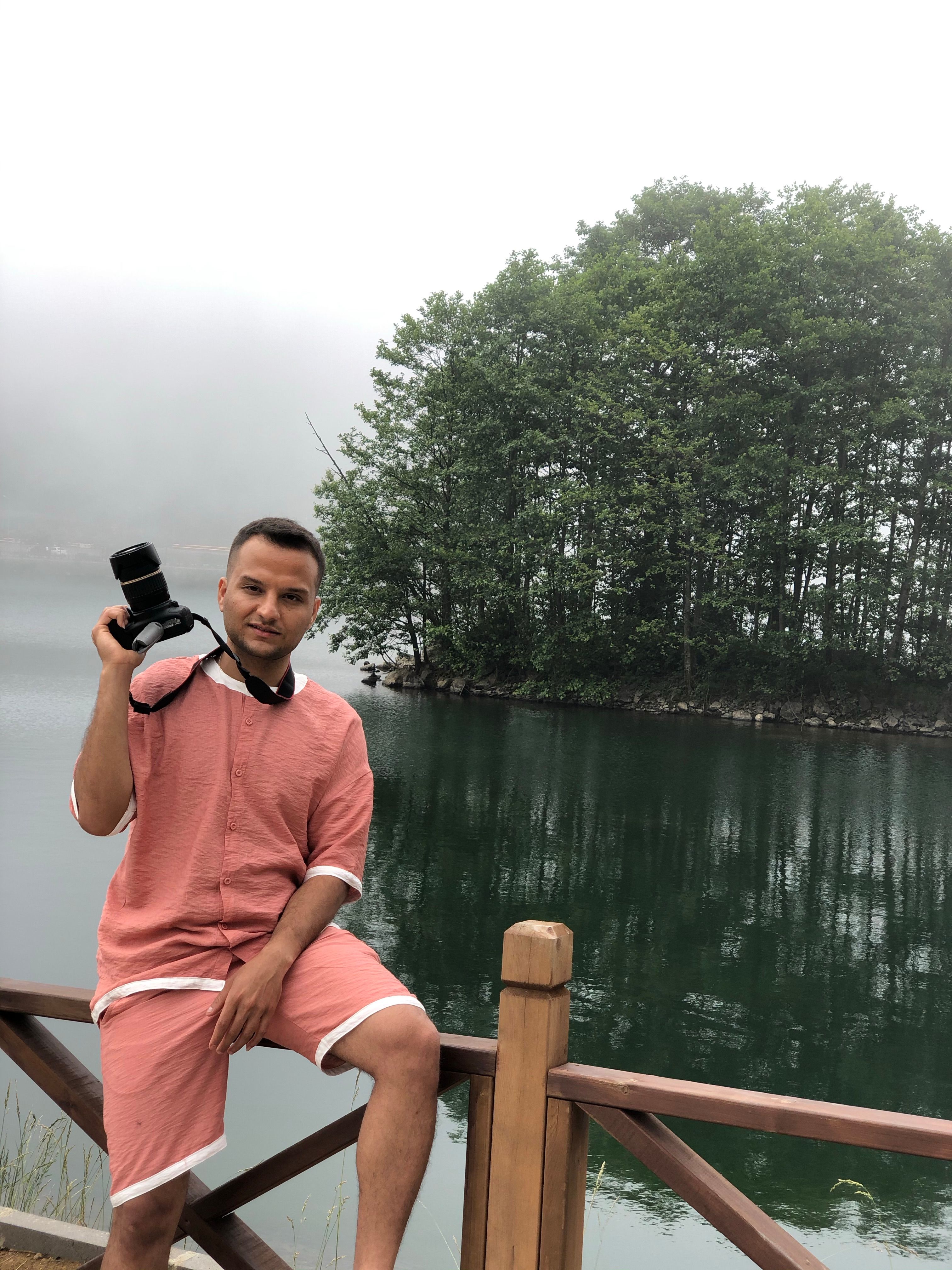 A man holding a camera sits by a tranquil lake surrounded by a misty forest, providing a serene outdoor scene.