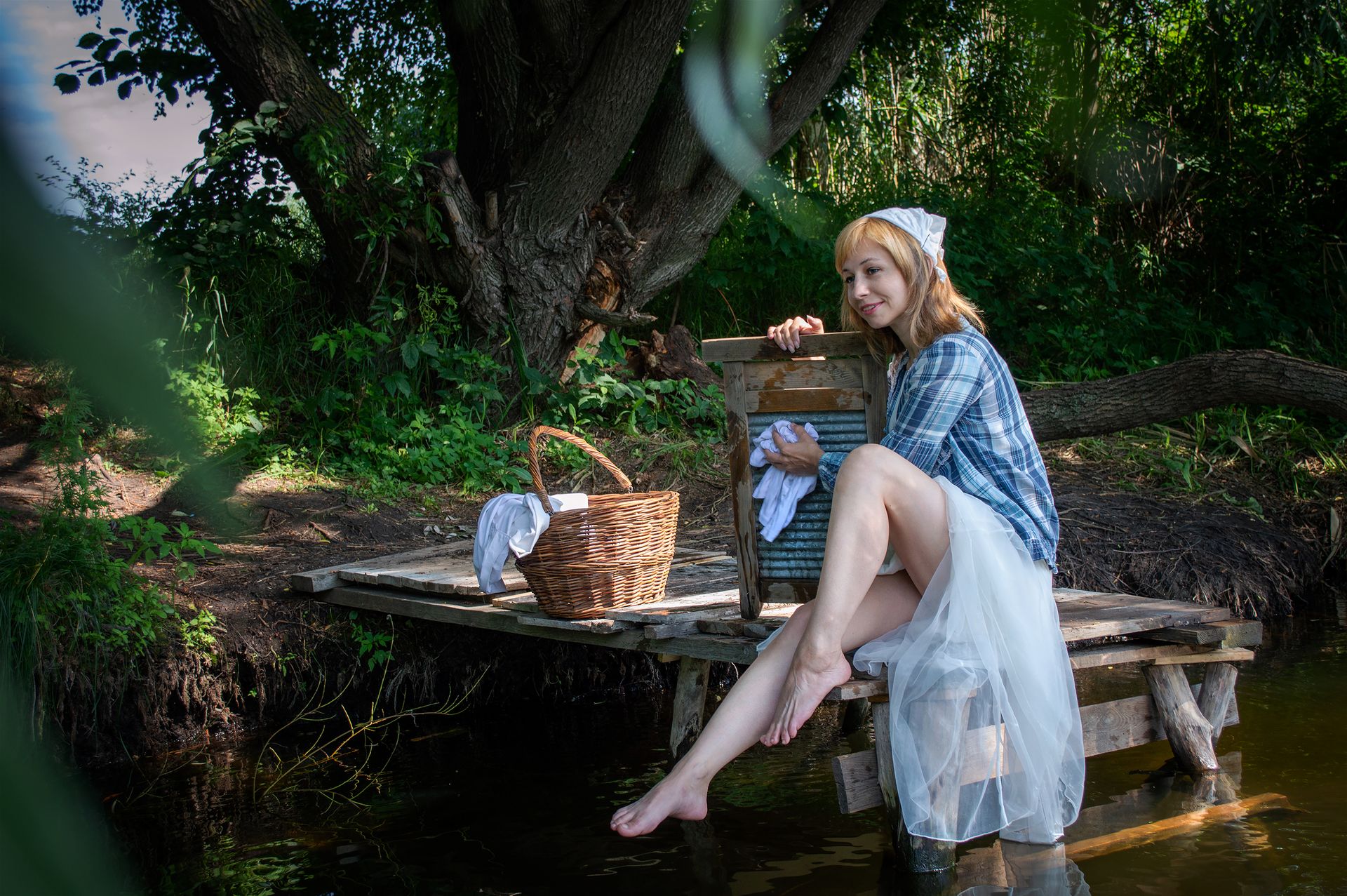 Woman sitting by a river doing laundry with a washboard, surrounded by nature.