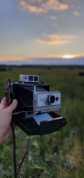 A hand holding a vintage analog camera with a blurred sunset background in a field.