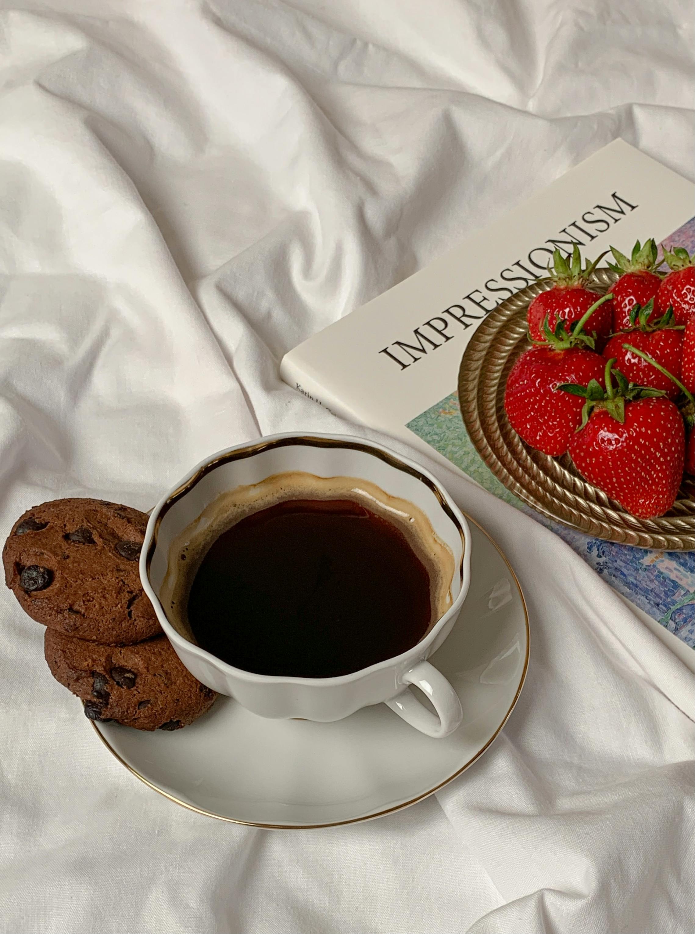 A cozy setup featuring coffee, cookies, strawberries, and an art book, perfect for a relaxing morning.