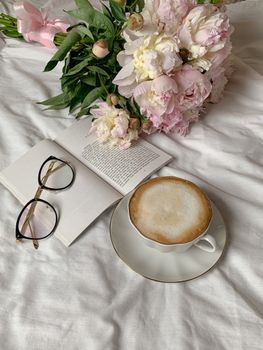 Elegant arrangement with pastel peonies, open book, and coffee on a bed for a relaxing vibe.