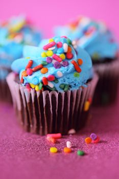 Delicious chocolate cupcakes topped with vibrant blue frosting and colorful sprinkles on a pink background.