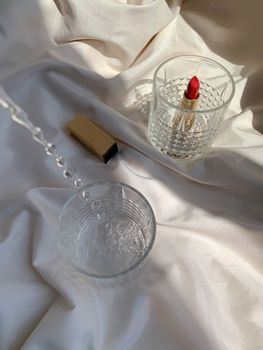 Aesthetic still life featuring red lipstick and crystal glasses on silky white fabric.
