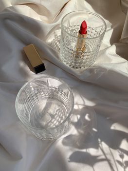 Aesthetic arrangement of lipstick and crystal glassware on satin fabric, capturing soft shadows.