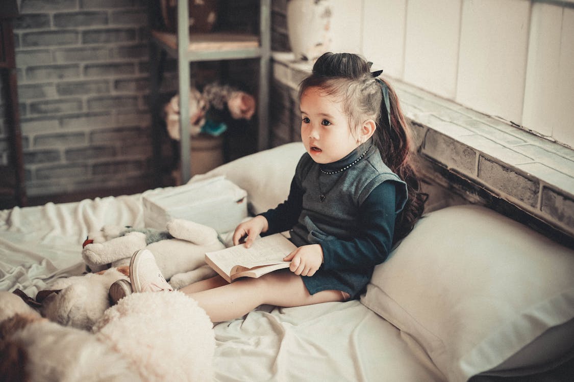 Free Toddler Girl Wearing Long-sleeved Top Reading Book While Sitting on Bed Stock Photo