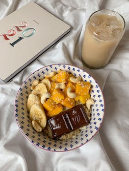 Delicious breakfast bowl with fruits, nuts, and chocolate, alongside iced coffee on white fabric.