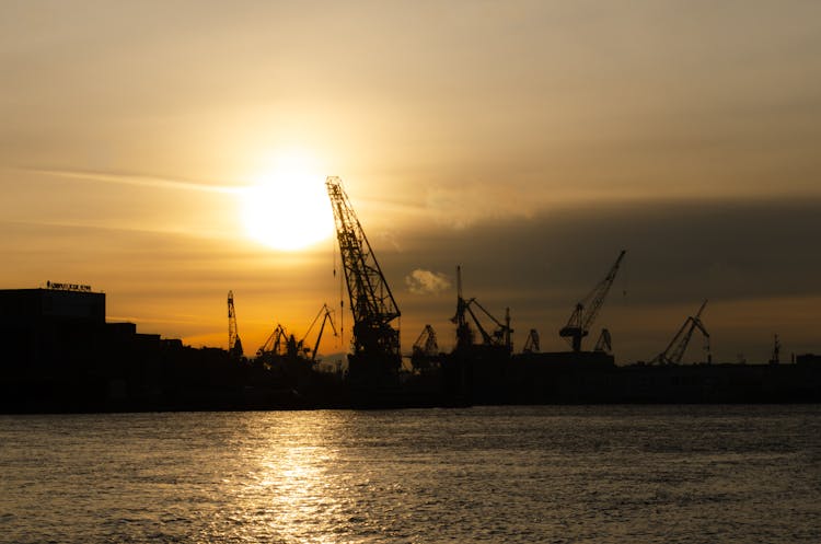 Silhouettes Of Shipyard Construction Cranes Against Sunset