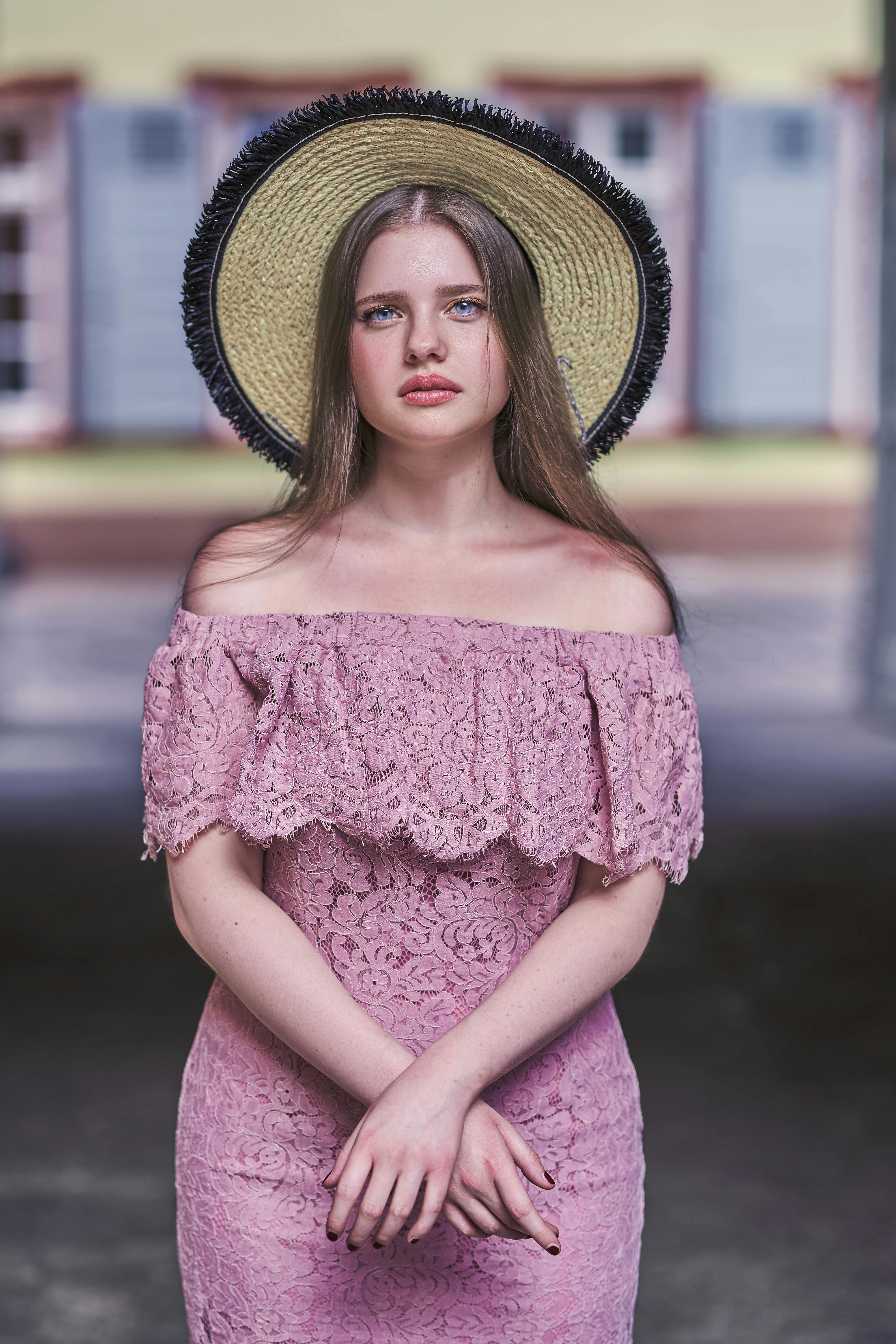 A woman in a pink lace dress with a sun hat poses elegantly outdoors against a blurred background.