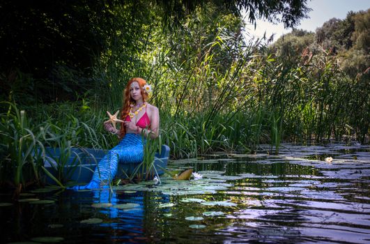 Stunning mermaid with red hair holds a starfish on riverbank, surrounded by lush nature.