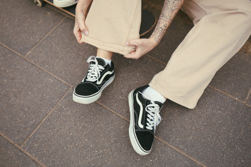 Free A person with a tattoo adjusts their beige pants, showcasing black sneakers on a paved surface. Stock Photo