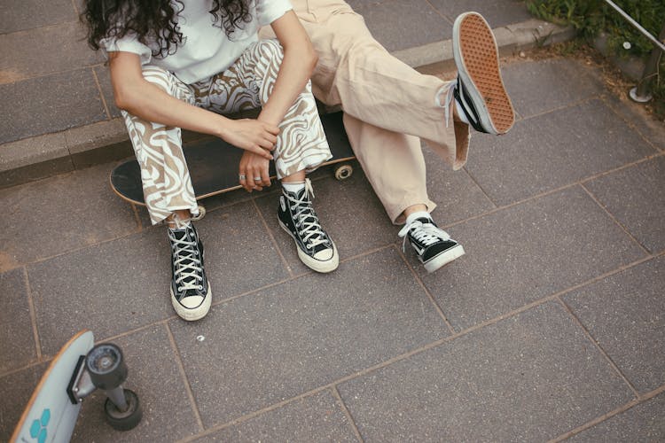Two Persons Wearing Pants And Sneakers Near A Skateboard