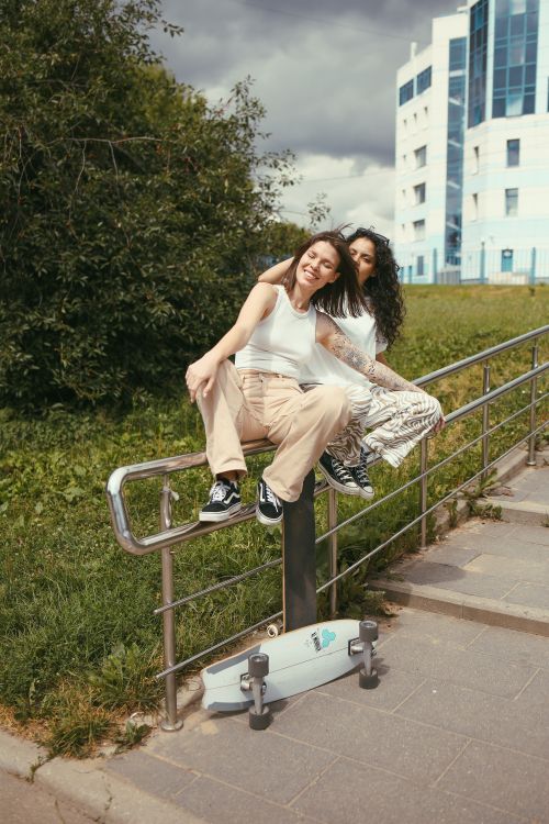Free Two happy young women sitting on a metal railing with a skateboard, enjoying a sunny day. Stock Photo