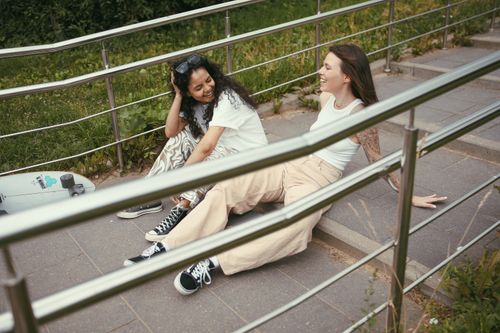 Free Two women enjoying a sunny day sitting on outdoor steps with a skateboard nearby. Stock Photo