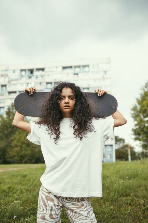 Free Confident woman skateboarder with curly hair poses in a city park, holding a skateboard above her shoulders. Stock Photo