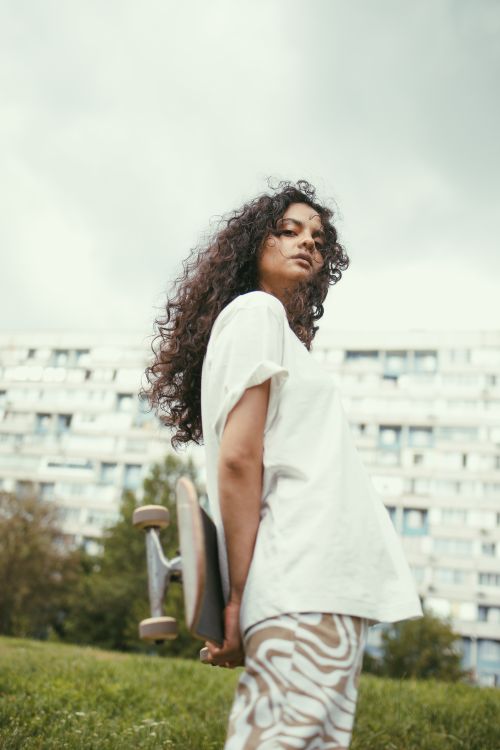 Free A young woman with curly hair holds a skateboard in an urban park with a city building backdrop. Stock Photo