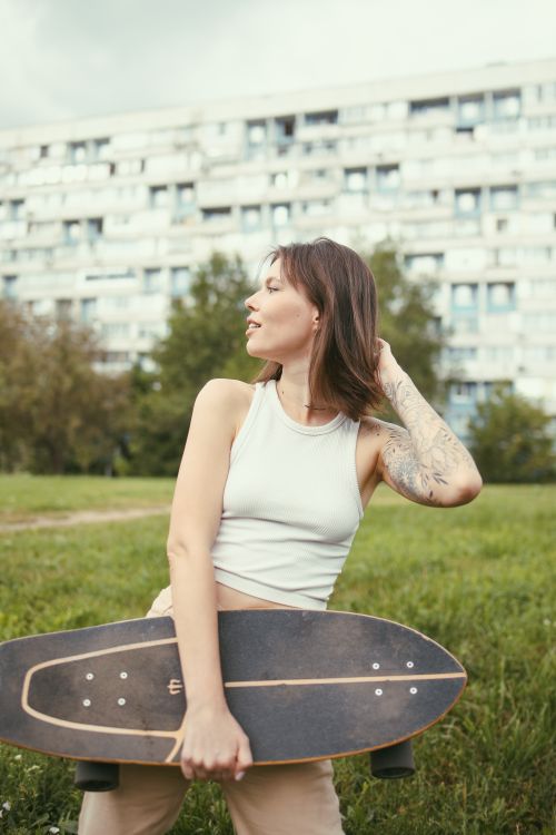 Free Portrait of a young woman holding a longboard in a green urban park with buildings in the background. Stock Photo