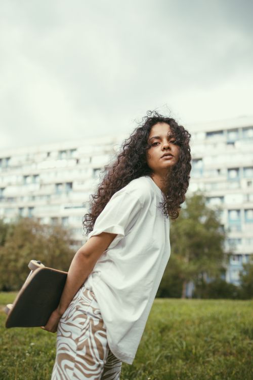 Free Confident young woman holding a skateboard in a park against a backdrop of urban buildings. Stock Photo