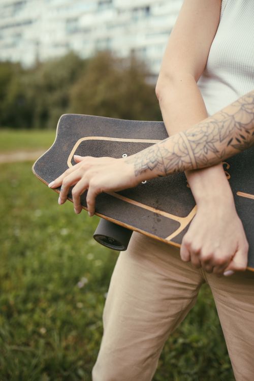 Free A tattooed person holds a longboard in a park, symbolizing outdoor activity and urban style. Stock Photo