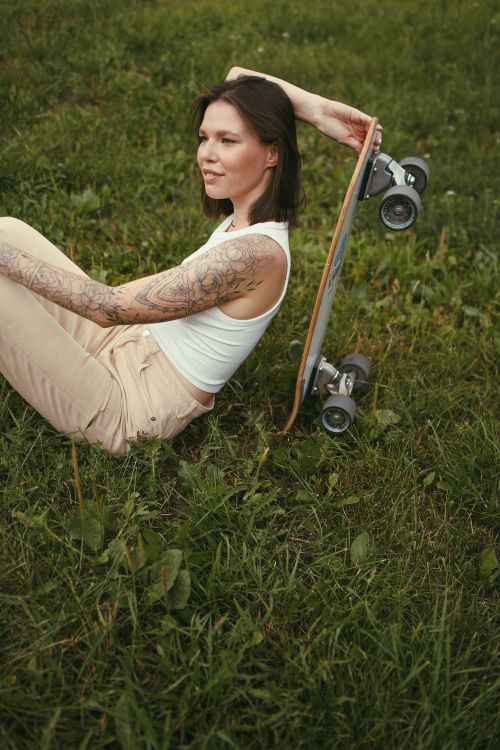Free Woman sitting outdoors with skateboard on grass, showing arm tattoos and enjoying a peaceful moment. Stock Photo