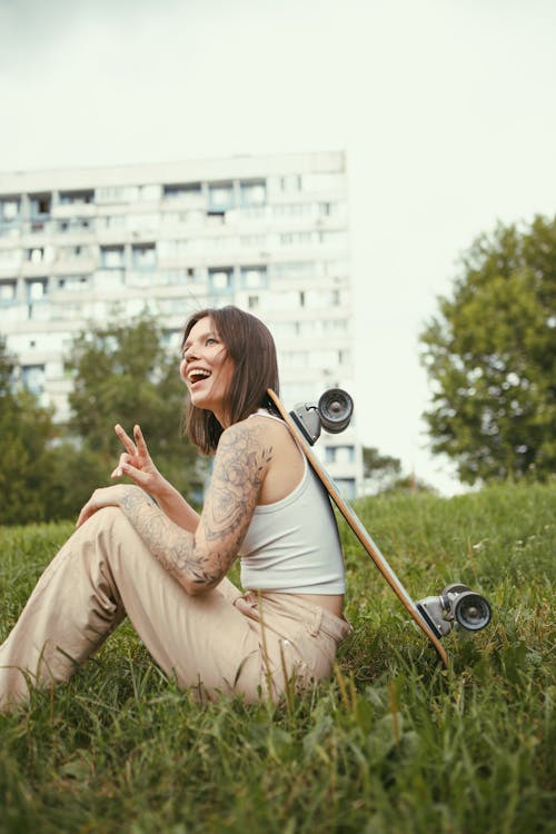 A Woman in White Tank Top Sitting on Green Grass with a Skateboard Leaning on her Back