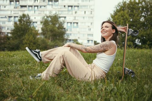 Free Young woman with tattoo sitting on green grass beside skateboard, urban setting. Stock Photo