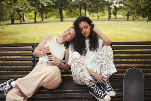 Free Two women enjoying a relaxing day, sitting on a bench outdoors with a skateboard nearby. Stock Photo