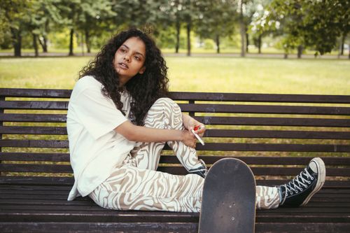 Free Woman with curly hair and skateboard sitting on a bench in a park, smoking. Stock Photo