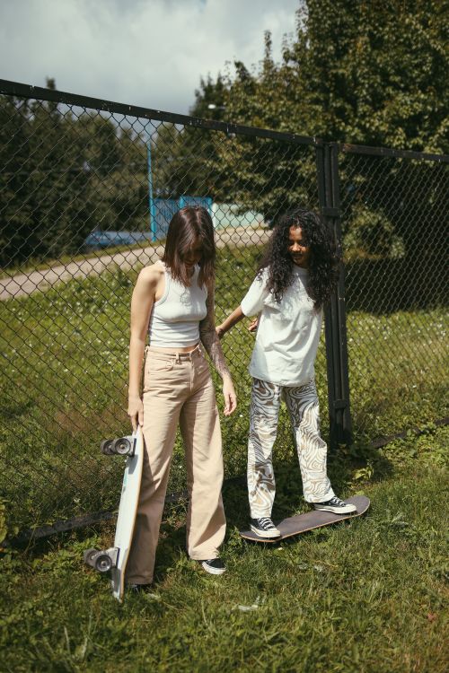 Free Two young women enjoying a day outdoors with skateboards, standing by a fence in a sunny park. Stock Photo