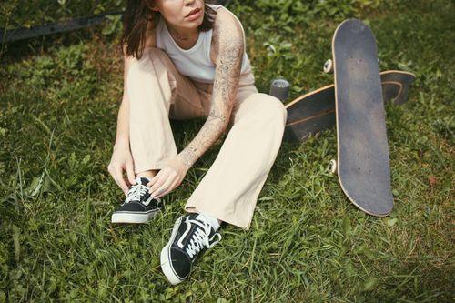 Free A woman with tattoos ties her shoes while sitting on grass with a skateboard nearby. Stock Photo