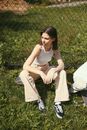 Woman in White Tank Top and White Pants Sitting on Green Grass