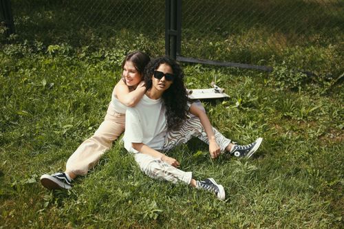 Free Two women sitting on grass near a fence, enjoying a sunny day with a skateboard. Stock Photo