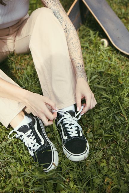 Close-up of a woman tying her black Vans sneakers in a park setting, showcasing casual style and tattoos.