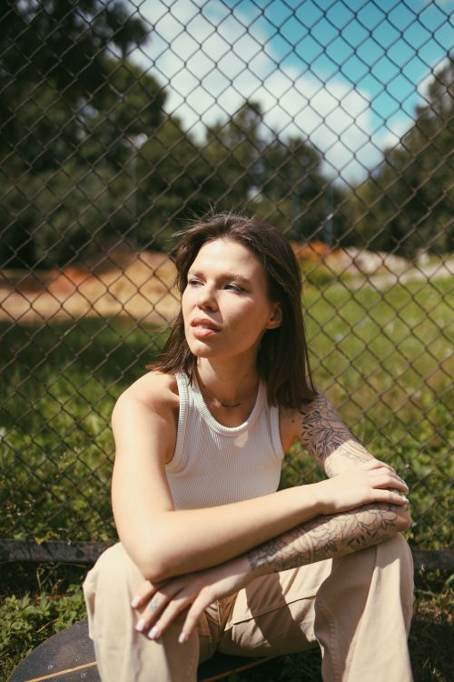 Free A young woman sits pensively by a chain link fence in a sunlit outdoor park. Stock Photo