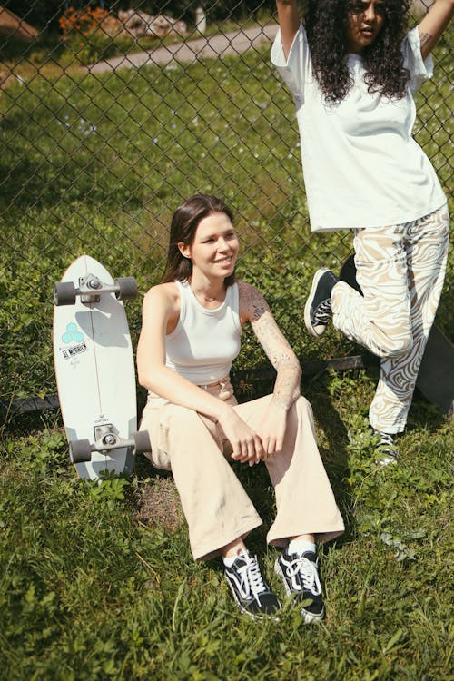 A Girl in White Tank Top and Beige Pants Sitting on Grass Beside a Skateboard