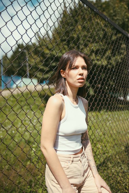 Free A young woman with short hair leans against a chain link fence outdoors in daylight. Stock Photo