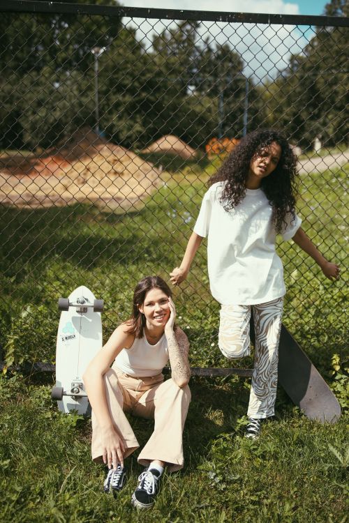 Free Two young women relaxing by a chain link fence with skateboards on a sunny day. Stock Photo