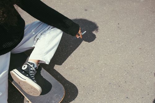 Free Person in black sweater with skateboard, sitting on concrete, holding a cigarette. Stock Photo