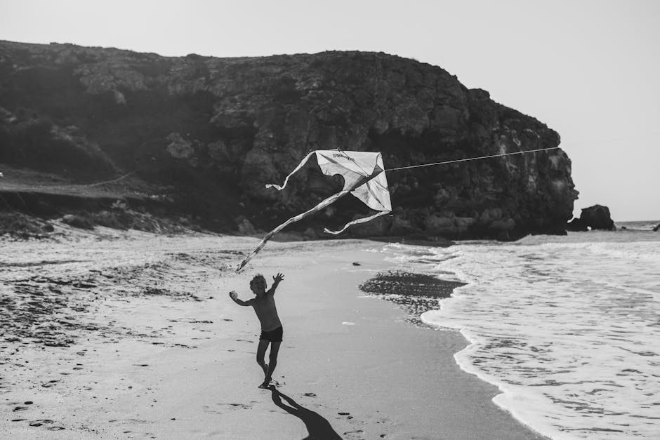 Black and white photo of a child flying a kite on a secluded beach.