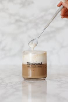 Cappuccino with creamy foam being stirred by a spoon, in a glass cup labeled 'Weekend'.