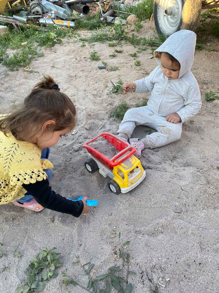 A Baby And A Girl Playing On Sand With A Toy Truck