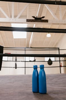 Blue reusable water bottles on the floor of a boxing gym with rings and bags in the background.